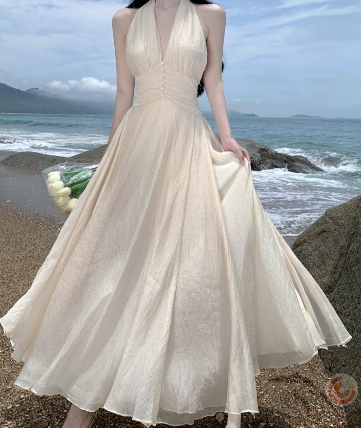 Woman in a flowing beige dress standing on a beach with ocean and sky in the background